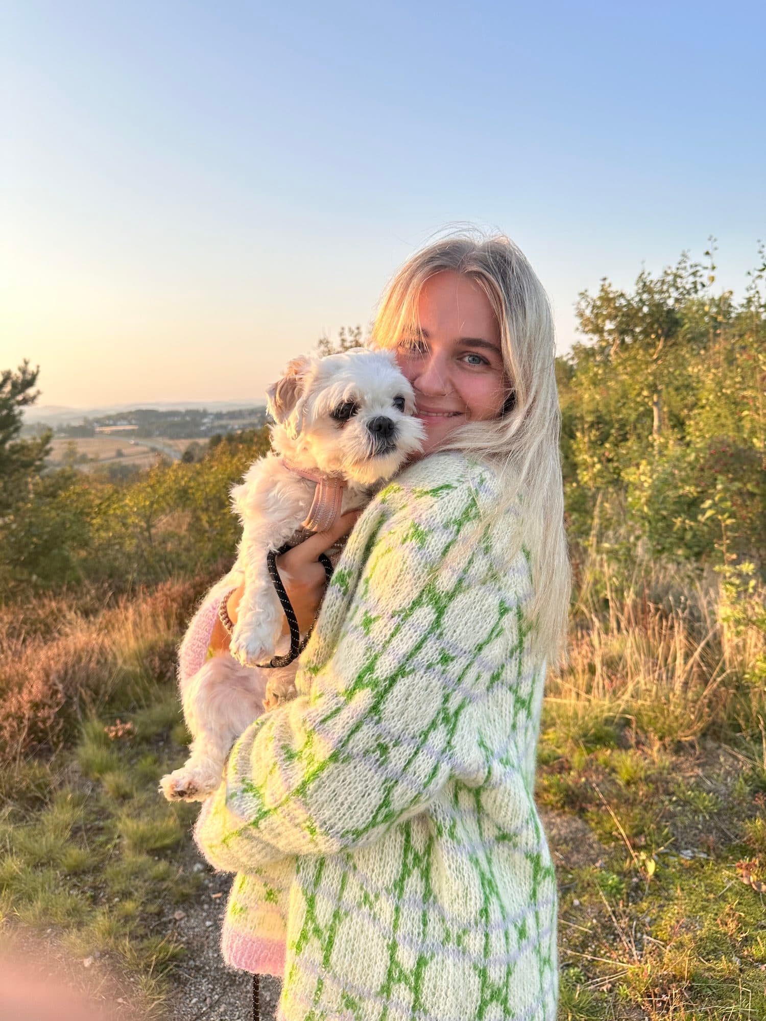 Frida i håndstrikket genser holder en liten hund utendørs i solnedgang
