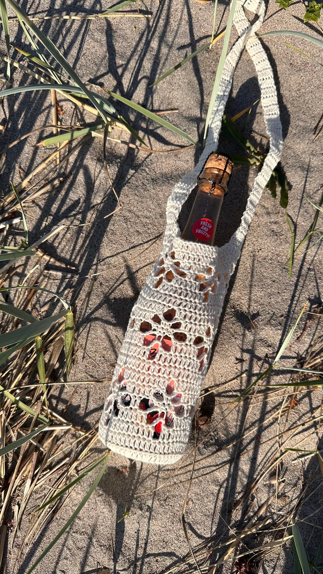 Heklet vinveske med flaske liggende i sanden på stranden